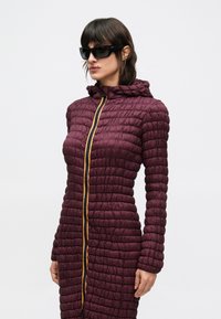 Gepolsterter bordeauxroter langer Mantel mit strukturierter Oberfläche, ausgestattet mit einem gelben Reißverschluss und Kapuze, getragen mit schwarzer Sonnenbrille.