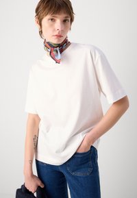 Vit bomulls t-shirt med rund hals, tillsammans med mörkblå denimjeans. En färgglad silkscarf är knuten runt halsen. Tatuering synlig på armen.