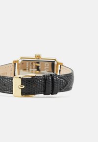 Schwarzes, strukturiertes Lederarmband mit goldfarbener Schnalle und Verschluss. Rechteckiges Zifferblatt mit reflektierender Rückseite und ohne sichtbare Markierungen.