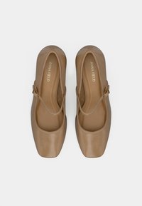 Svetlé hnedé Mary Jane balerínky so štvorcovými špičkami, tenkými pásikmi a zlatými prackami na bielom pozadí.