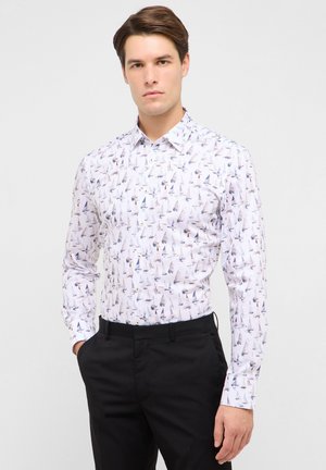 Uomo con capelli castani che indossa una camicia bianca con piccoli motivi di barche a vela multicolori e pantaloni neri, in piedi con le mani nelle tasche.