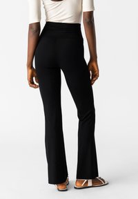 Zwarte flared broeken van rekbaar materiaal, met een hoge tailleband en een gladde textuur. Gecombineerd met platte, sandalen met straps.