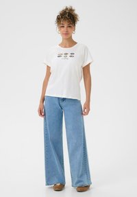 Frau steht da und trägt ein weißes T-Shirt mit Tierdruckmustern und hellblauen weit geschnittenen Jeans sowie braunen Schuhen.