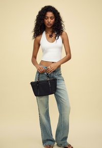 Borsa a tracolla nera con manici corti, chiusura con zip e dettagli metallici. Il modello indossa un crop top bianco e jeans a zampa d'elefante blu chiaro su uno sfondo beige.