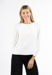 Blusa bianca con dettagli a balze sulle maniche, caratterizzata da un colletto rotondo e un motivo testurizzato discreto. Abbinata a pantaloni neri.