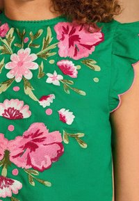 Grön topp med intrikata blommönster i rosa, vitt och rött. Vågmönstrade ärmslut och mjuk tygtextur är synliga.