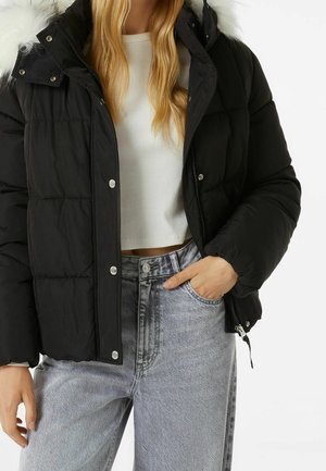 Femme portant une doudoune noire avec une capuche doublée de fourrure, un pull court blanc et un jean taille haute délavé clair avec la main dans la poche.