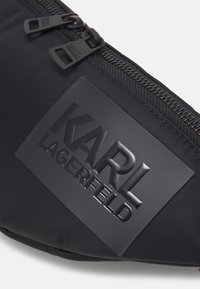 Černé nylonové bum bag s dvojitými zipy, texturovanou nášivkou loga a hladkým povrchovým zpracováním. Kompaktní design bez viditelných kovových detailů.