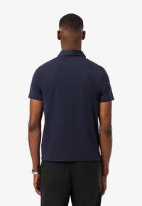 Hombre con camiseta tipo polo de manga corta azul marino y pantalones negros, de pie de espaldas al espectador contra un fondo blanco.