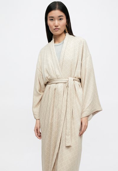 Κρεμ ρόμπα σε στιλ κιμονό με διαμαντένιο μοτίβο, με ζώνη στη μέση. Διαθέτει φαρδιά μανίκια και απαλό υφή, ντυμένη πάνω από ένα γκρι μπλουζάκι.
