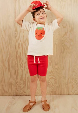 Ein Kind trägt ein cremefarbenes T-Shirt mit einem Erdbeer-Graphic und dem Text "BERRY KIND", kombiniert mit roten Shorts und braunen Sandalen vor einem Holzhintergrund.