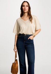 Beige, løs strikket top med V-hals og korte ærmer, parret med højlivet mørke jeans. Holder en tan fringet håndtaske.