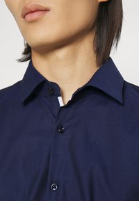 Námořnicky modrá košile na knoflíky z texturované látky, s rozšířeným límcem a kontrastní bílou vnitřní légou. Černé knoflíky vpředu.