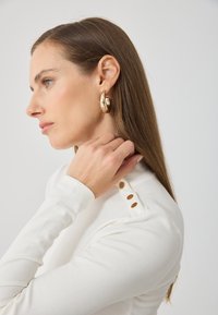 Biele pruhované tričko s dlhým rukávom má na ramene tri zlaté gombíky. Modelka nosí lesklé zlaté náušnice vo forme kruhu a dotýka sa svojho krku.