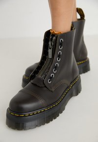 Botas de tobillo de cuero marrón con una suela negra gruesa, costuras amarillas, cremallera frontal y adornos de ojales, con un acabado texturizado.