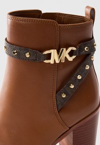 Brown leather ankle boot with dark studded strap and gold MK logo buckle, featuring a medium wooden block heel.
