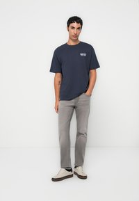 Camiseta de algodón azul marino con un pequeño logotipo blanco en el lado izquierdo del pecho, combinada con jeans rectos de color gris claro y zapatillas blancas.