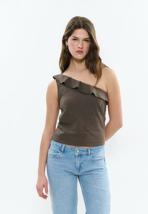 Jeune femme aux longs cheveux bruns portant un haut marron à volants, sans manches et à une épaule, ainsi qu'un jean bleu clair, debout devant un fond blanc.