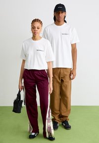 Zwei Modelle stehen zusammen. Eines trägt ein weißes T-Shirt und burgunderfarbene Sporthosen mit weißen Streifen; das andere hat ein weißes T-Shirt und braune Hosen an.