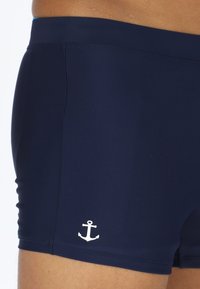 Shorts de bain navy en matériau extensible, présentant un emblème d'ancre blanche sur le côté gauche et un design ajusté.