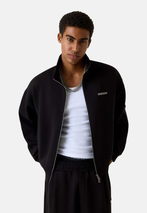 Giovane uomo con capelli corti e ricci, indossa una giacca nera con cerniera sopra una canottiera bianca e pantaloni neri, in piedi davanti a uno sfondo semplice.