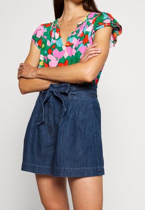Femme portant un short en jean taille haute avec une ceinture nouée et un chemisier à manches courtes à fleurs de couleurs verte, rose, rouge et bleue.