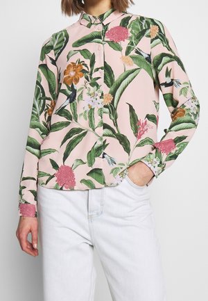 Femme portant une chemise rose pâle boutonnée avec des feuilles vertes, des fleurs colorées et des motifs d’oiseaux, associée à un jean bleu clair.