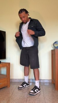 Giacca di jeans scura sopra una maglietta bianca, pantaloni corti grigi e scarpe da ginnastica nere Nike. Calzini bianchi visibili. In piedi su un pavimento piastrellato.