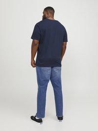 Camiseta de algodón azul marino de manga corta, vista trasera, combinada con jeans de mezclilla azul claro y zapatillas negras. Diseño simple y casual.