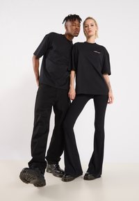 Two models wear black oversized T-shirts, paired with fitted black ribbed pants and black chunky boots, set against a plain background.