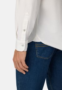 Camicia bianca a maniche lunghe con polsini a bottoni, caratterizzata da una texture liscia. Abbinata a jeans in denim blu scuro con dettagli di cucitura gialla.