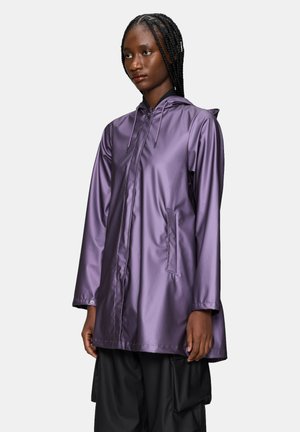 Donna con i capelli intrecciati che indossa un impermeabile viola lucido con cappuccio e pantaloni cargo neri a gamba larga, in piedi davanti a uno sfondo semplice.