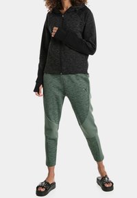 Chaqueta negra con cremallera y mangas texturizadas, combinada con pantalones joggers ajustados verdes con detalles en panel y costuras de acento; llevados con sandalias negras.