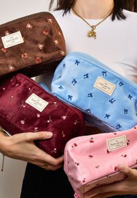 Cuatro bolsas de cosméticos de terciopelo en varios colores: marrón con abejas, burdeos con flores, azul con lazos y rosa con cerezas. Cierres de cremallera visibles.