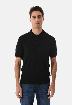 Uomo che indossa una polo nera a maniche corte e jeans blu, in piedi davanti a uno sfondo semplice grigio chiaro.