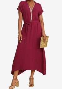 Burgundin pitkä mekko, jossa on lyhyet hihat, V-mallinen kaula-aukko ja kultaiset yksityiskohdat. Sidottu vyötärö ja korkea-alasolmuke. Yhdistetty tekstuuriseen kultaklutchin.