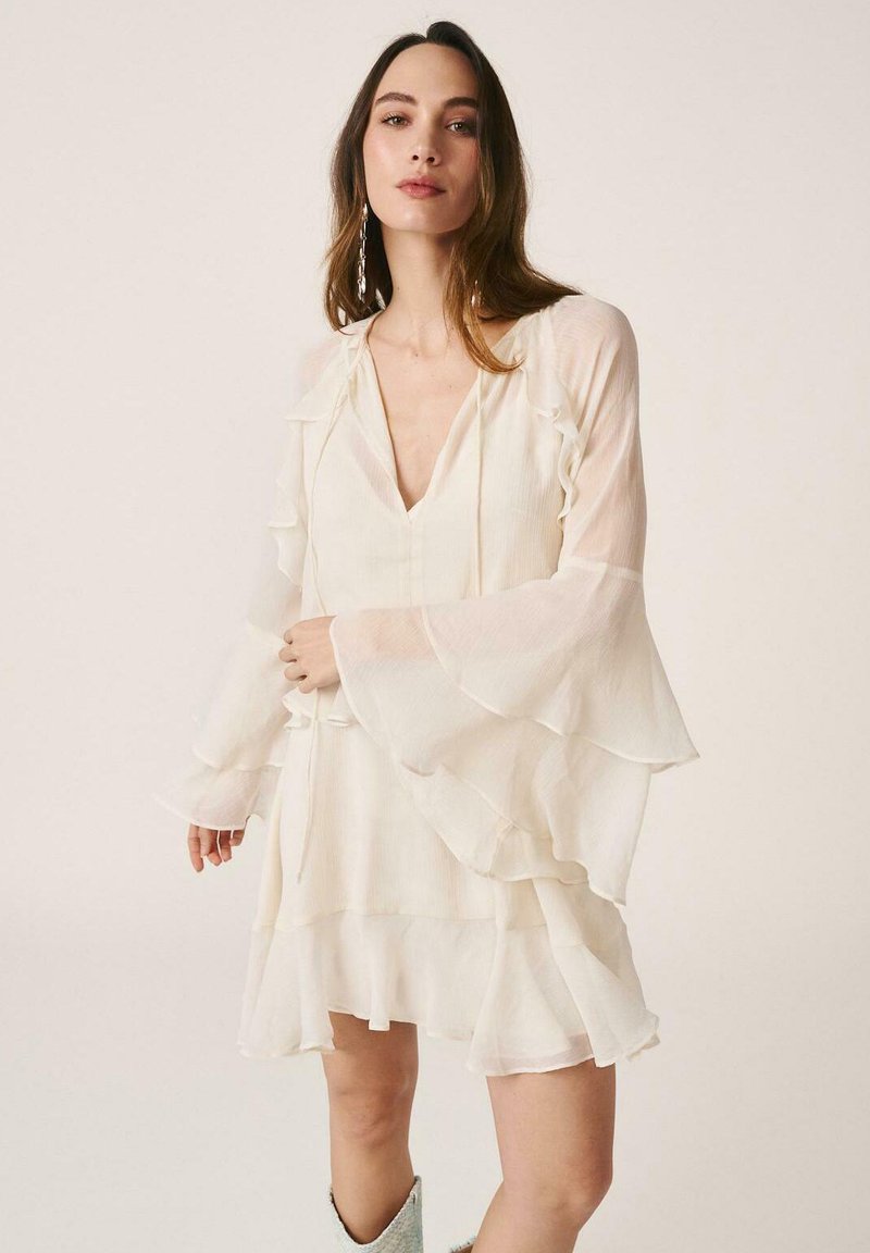 Mujer con un vestido blanco translúcido de capas, con mangas acampanadas y escote en V, de pie frente a un fondo claro y liso.