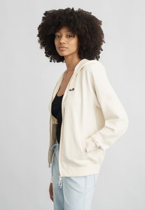 Νεαρή γυναίκα με σγουρά μαλλιά, φορώντας μια κρεμ φούτερ ζακέτα με φερμουάρ πάνω από μαύρο τοπ και ανοιχτό μπλε τζιν, μπροστά σε απλό φόντο.