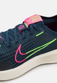 Marinblå atletiska skor med texturerad ovandel, rosa och gröna accenter samt Nike-logotyp. Innehåller en QR-kod och förstärkt tårområde.