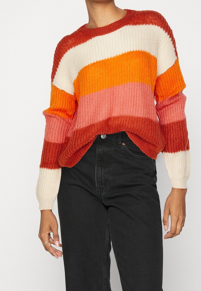 Gebreide trui met horizontale strepen in roest, wit, oranje en roze, gedragen met hoge zwarte jeans, handen ontspannen langs de zij.