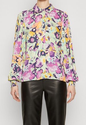 Vrouw die een pastelbloemenblouse met lange mouwen en gerimpelde manchetten draagt, en zwarte leren broek, staand tegen een effen achtergrond.