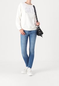 Felpa bianca con "GAP" in borchie argentate, abbinata a jeans skinny blu e sneaker bianche. Gli accessori includono una borsa nera con occhielli.
