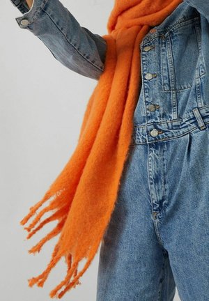 Veste en denim et jean taille haute portés avec une écharpe douce et brillante orange à franges sur un fond uni.