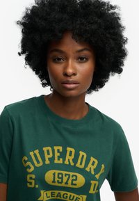 Grünes Baumwoll-T-Shirt mit Rundhalsausschnitt, großem gelben "SUPERDRY"-Schriftzug und einem grafischen Element, glatter Textur und lässigem Design.