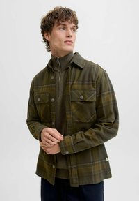 Olivgrüne karierten Hemdjacke mit zwei Brusttaschen, Knopfverschluss und einer strukturierten Stoffqualität. Getragen über einem dunklen Zip-Kragen-Pullover.