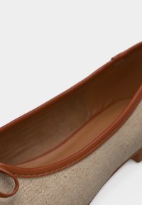 Μπεζ ύφασμα επίπεδη παπούτσια με καφέ δερμάτινη τελειώματα και μικρό τακούνι, εμφανίζεται από γωνιακή κορυφή.