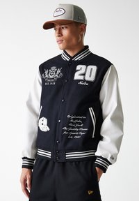 Black and white varsity jacket with embroidered details, ribbed collar, button closure, and wide sleeves. Pairing with a beige cap.