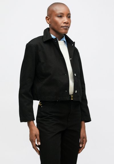 Isik, kellel on habemeajatud pea, kannab musta lühikest jakki, valget kampsunit, helesinist särki ja musti pükse, seistes valge tausta ees.