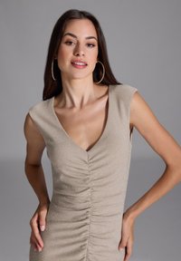 Polvepituinen, tyköistuva mekko metallisen beigen värisenä, jossa on lyhyet hihat, syvä V-pääkallo ja rypytetty rakenne edessä.