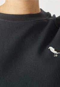 Felpa blu scuro con scollatura rotonda, caratterizzata da un piccolo design di uccello ricamato in bianco e marrone sulla zona del petto.